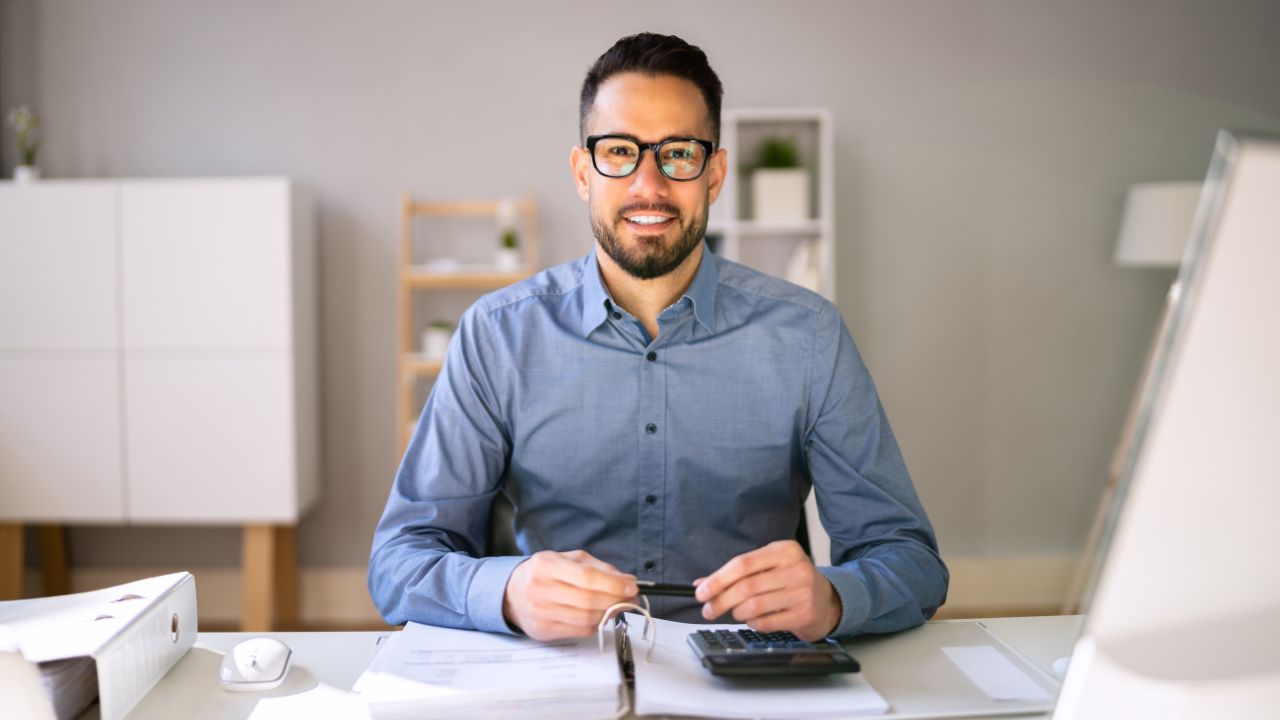 Smiling man in glasses sitting at a desk with a calculator and paperwork